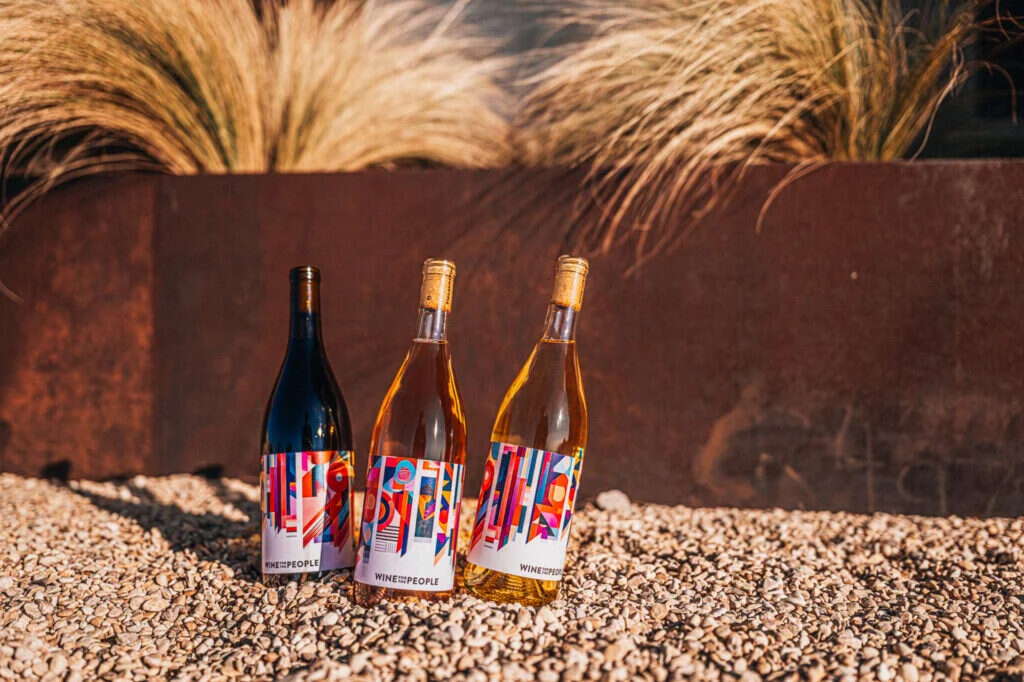 Three Wine For The People bottles with colorful abstract labels on a gravel surface