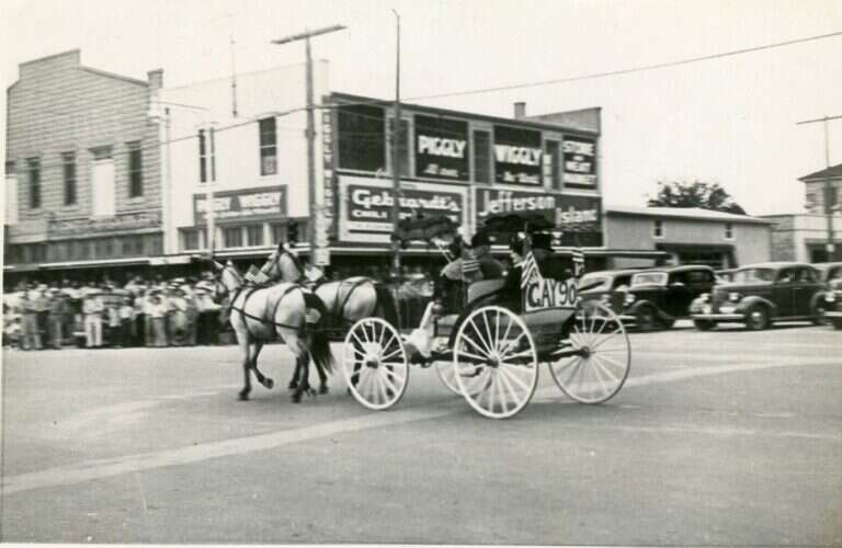 Horse-drawn carriage in a parade on Main Street in Fredericksburg, Texas, passing painted Piggly Wiggly signs on the side of the Maier Building