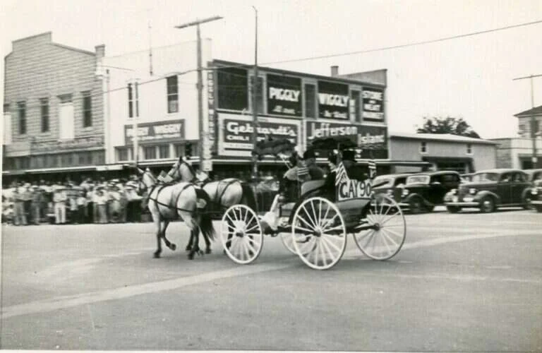 Horse-drawn carriage in a parade on Main Street in Fredericksburg, Texas, passing painted Piggly Wiggly signs on the side of the Maier Building