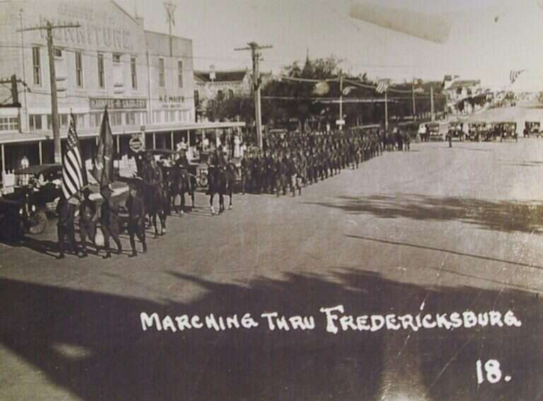 Soldiers and mounted riders marching down Main Street in Fredericksburg, Texas, past H.C. Maier's store and its harness and saddlery sign