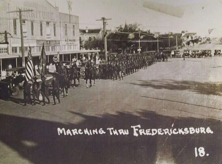 Soldiers and mounted riders marching down Main Street in Fredericksburg, Texas, past H.C. Maier's store and its harness and saddlery sign