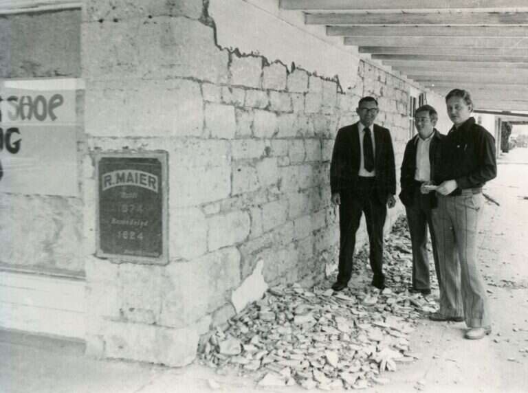Three men stand beside exposed limestone and an R. Maier plaque reading Built 1874, Remodeled 1924, on the Maier Building in Fredericksburg