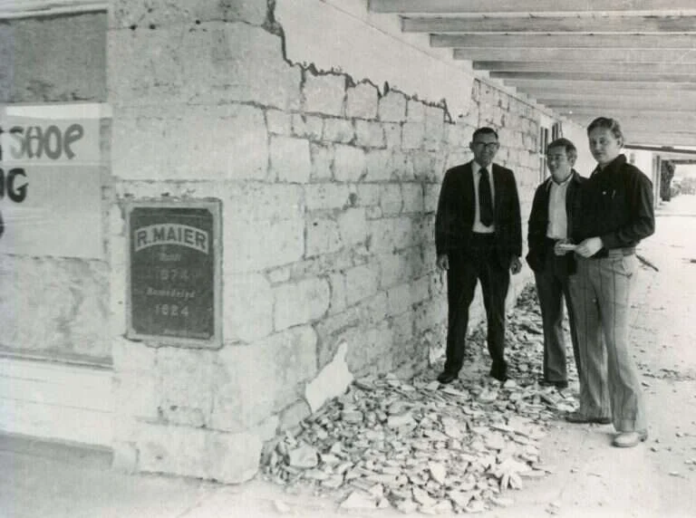 Three men stand beside exposed limestone and an R. Maier plaque reading Built 1874, Remodeled 1924, on the Maier Building in Fredericksburg