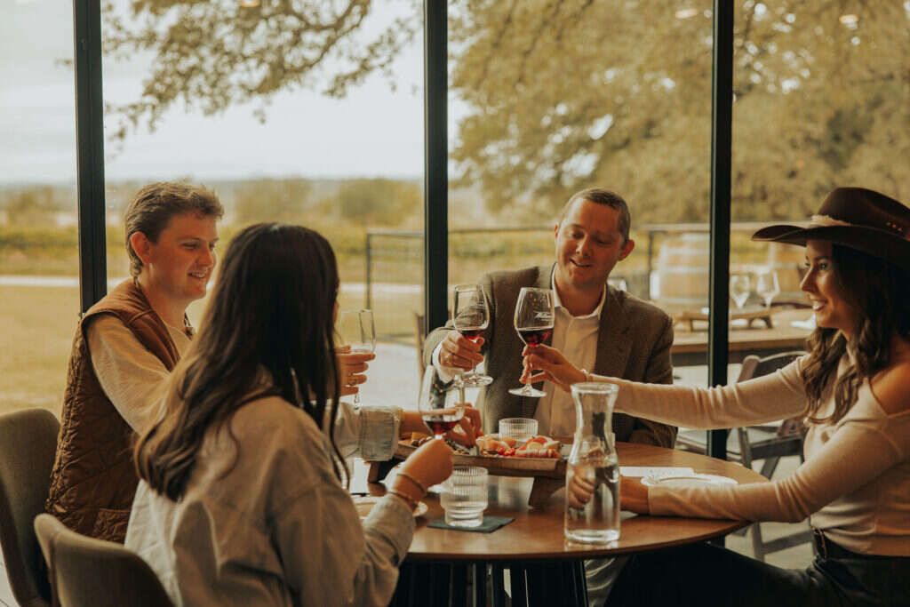 Four friends toasting with glasses of red wine at a table in the William Chris tasting room in Hye, with the vineyard through the windows behind them